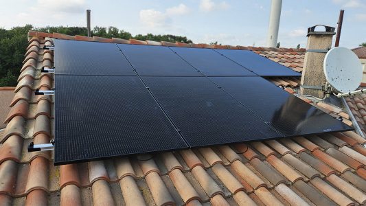 Panneaux solaires noirs installés sur un toit de tuiles en terre cuite, présence d'une antenne satellite et d'une cheminée.