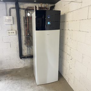 Une pompe à chaleur Daikin dans une cave, avec des tuyaux et un mur en blocs de béton blancs.