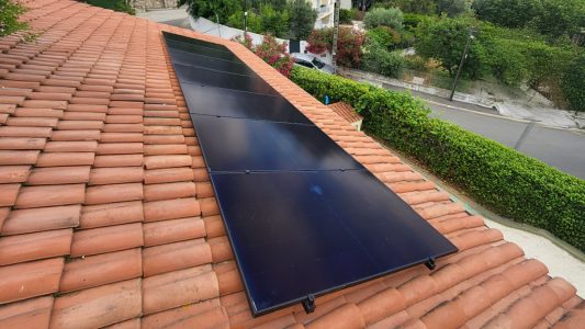 Panneaux solaires noirs installés sur un toit en tuiles rouges, entourés d'arbres et d'une rue.