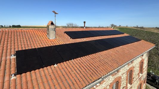 Panneaux solaires sur le toit d'une maison en briques, avec un paysage rural en arrière-plan et un ciel bleu clair.