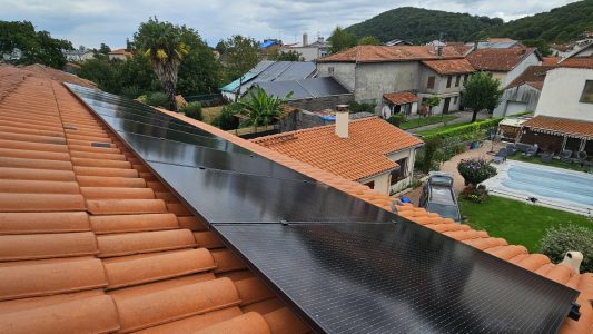 Panneaux solaires noirs installés sur un toit en tuiles rouges dans un paysage verdoyant, avec des maisons en arrière-plan.