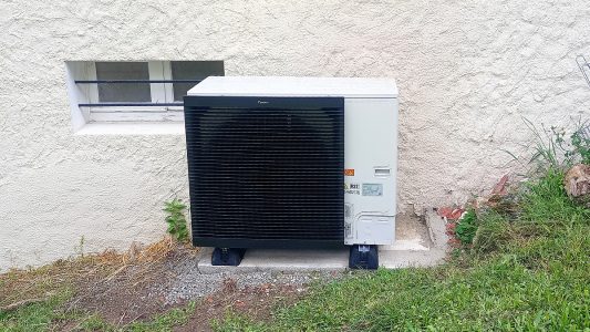 Climatiseur extérieur Daikin, installé contre un mur blanc texturé, près d'une petite fenêtre et de la végétation.