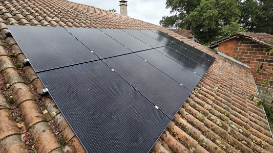 Panneaux solaires noirs sur un toit en tuiles avec un arrière-plan de verdure et de ciel nuageux.